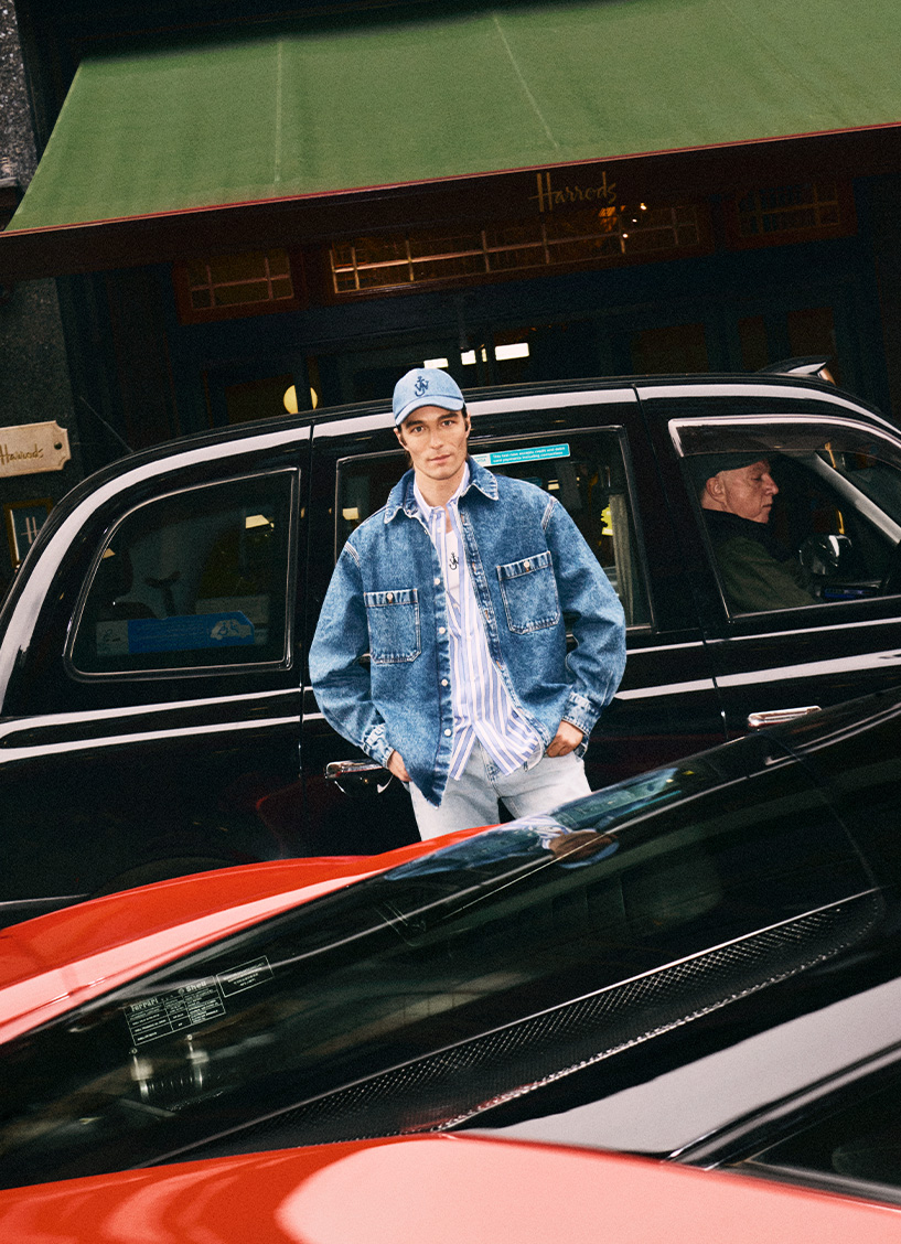 Wearing a blue denim jacket and cap, a male model stands in front of a London taxi which is parked in front of Harrods