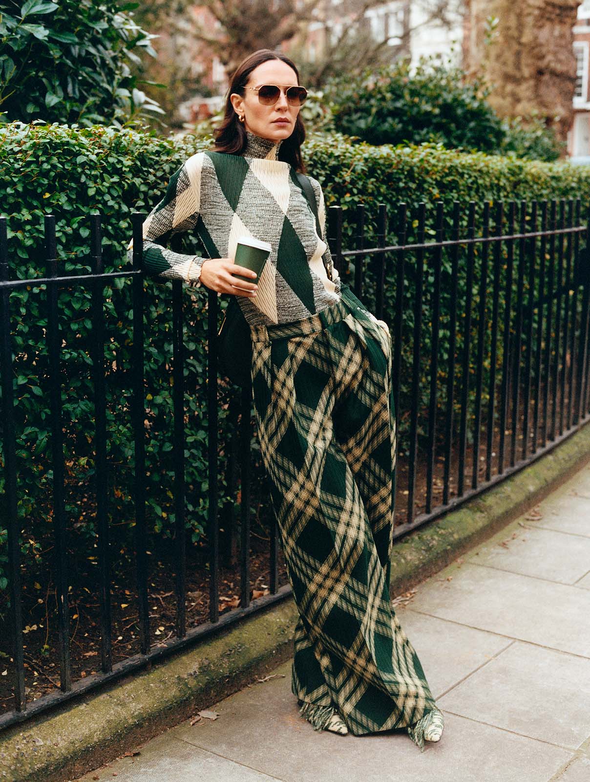 Woman wears green check Burberry jumper and trousers, holding a Harrods takeaway coffee cup